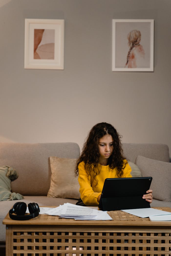 Teen girl focused on laptop at cozy living room workspace with headphones and papers around.
